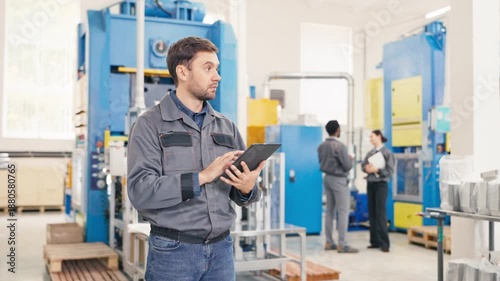 Caucasian male worker using digital tablet entering production data. Focused expression while reviewing information. Two colleagues discussing tasks in blurred background. Bright industrial hall.