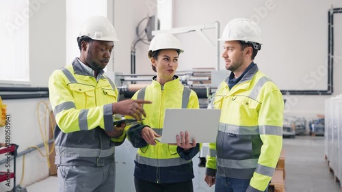 Group of factory engineers wearing safety jackets and helmets discussing project. Female holding laptop showing data while colleague using tablet. Team collaborating in production facility.
