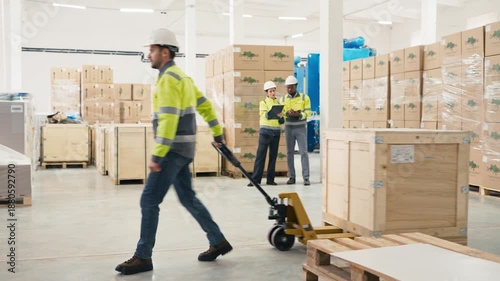 Factory male worker pulling loaded pallet truck across warehouse floor. Colleagues in reflective uniforms reviewing documents in background. Busy day. People surrounded by boxes and packed goods.