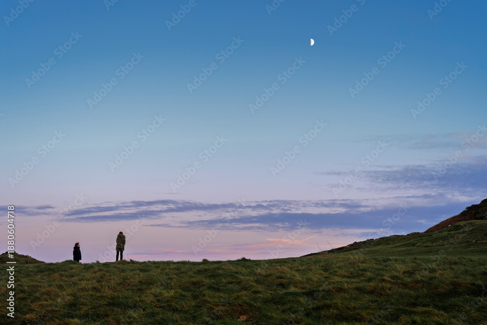 Fototapeta premium Wicklow Head dusk with two silhouetted figures, textured grass and half-moon over serene Irish Sea, autumn evening light