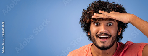 A young man stands with his hand over his forehead, looking ahead with a big smile. He has curly hair and is wearing a casual shirt. The background is blue, which adds to the cheerful mood.