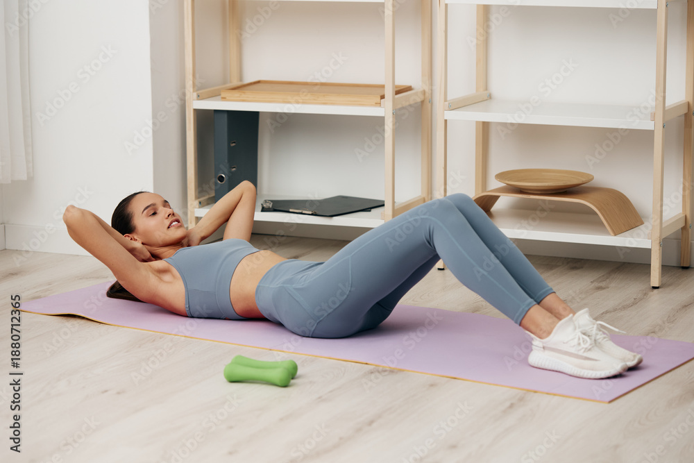 Obraz premium Woman fitness, sport scene: young woman training at home performing crunches on a yoga mat in comfortable activewear, focused expression, clean minimalist room and a small dumbbell nearby.