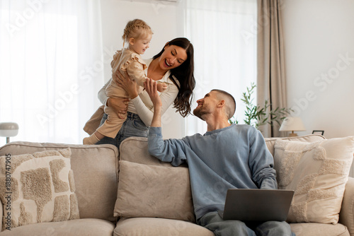 Tablou pe pânză Mother carrying infant daughter father using laptop, working remotely from home