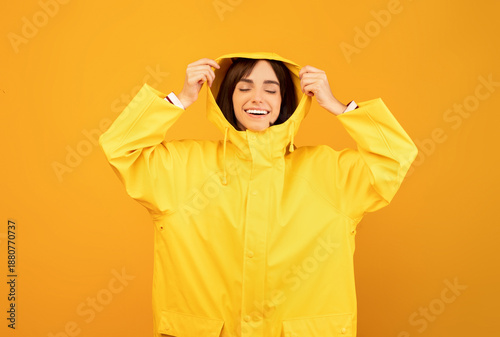Fotografija Excited millennial lady in yellow waterproof raincoat putting on hood, standing over yellow background and smiling with closed eyes