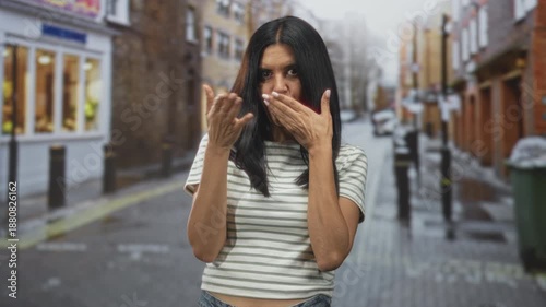 Woman with hand covering mouth and other hand making a beckon gesture on a wet cobbled street; playful flirtation.