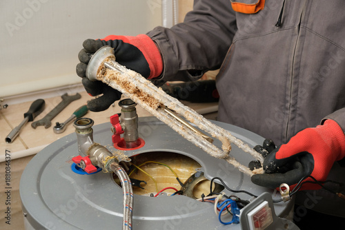 Photos Repairman in red gloves demonstrates a burnt-out boiler heating element covered with lime coating