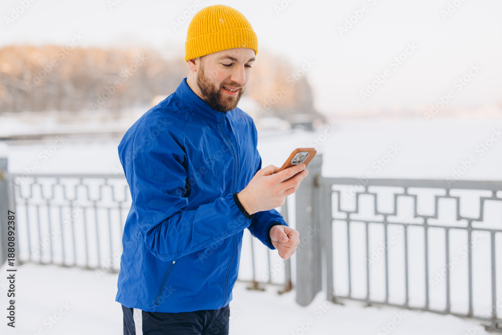 Obraz premium Male runner checking workout data on smartphone during a cold winter outdoor training session in snow