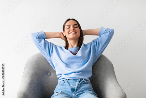 Принти на полотні Pretty young Caucasian woman sitting in comfy armchair with hands behind her head, closing eyes, relaxing on white studio background