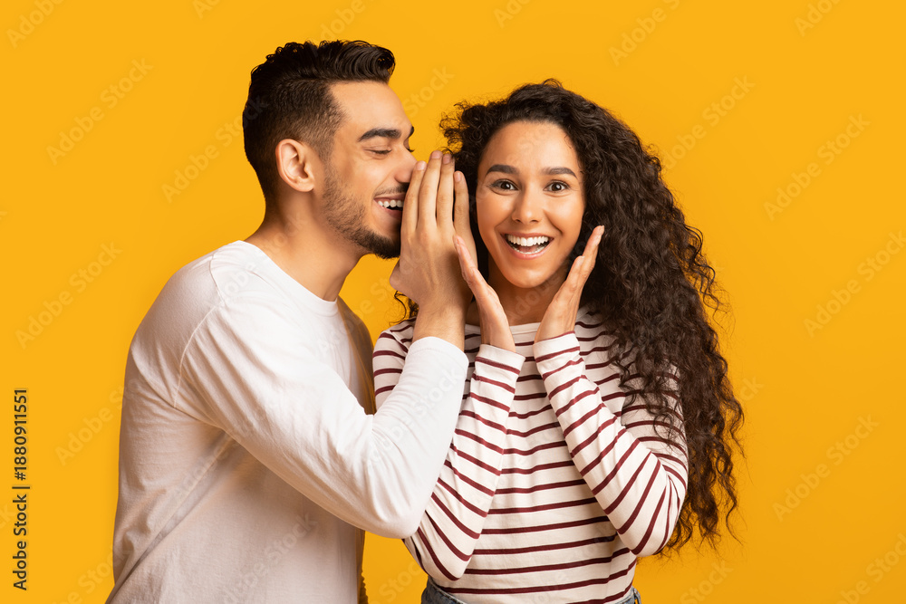 Naklejka premium Big Secret. Young Arab Man Sharing News With His Excited Girlfriend, Surprised Middle Eastern Woman Raising Hands In Amazement While Standing Together Over Yellow Studio Background, Free Space