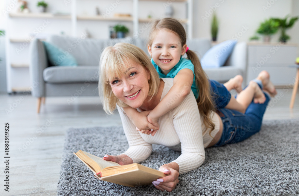 Naklejka premium Story Time. Portrait of excited mature woman and cute little girl reading book together lying on rug floor carpet in living room, looking at camera, staying at home, free copy space