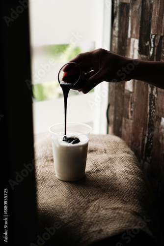 Fotografi Coffee syrup poured into milk in plastic cup on burlap fabric, moody cafe scene for beverage preparation, coffee shop branding, and advertising use