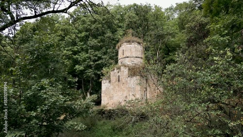 Drone panoramic flyover of a medieval stone Christian church surrounded by mountains and dense forest. Trees grow on the church roof. No people, only nature. Cinematic aerial shot.