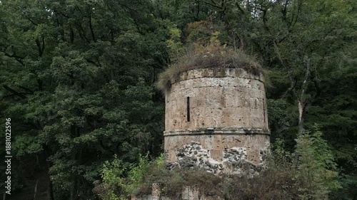 Drone panoramic flyover of a medieval stone Christian church surrounded by mountains and dense forest. Trees grow on the church roof. No people, only nature. Cinematic aerial shot.