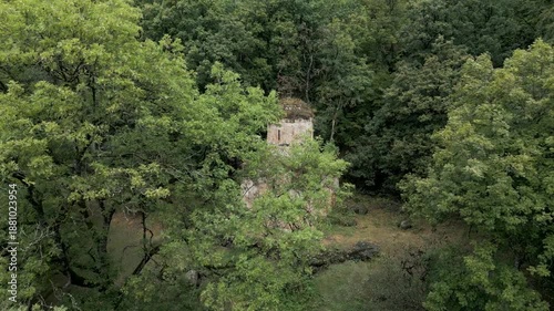 Drone panoramic flyover of a medieval stone Christian church surrounded by mountains and dense forest. Trees grow on the church roof. No people, only nature. Cinematic aerial shot.