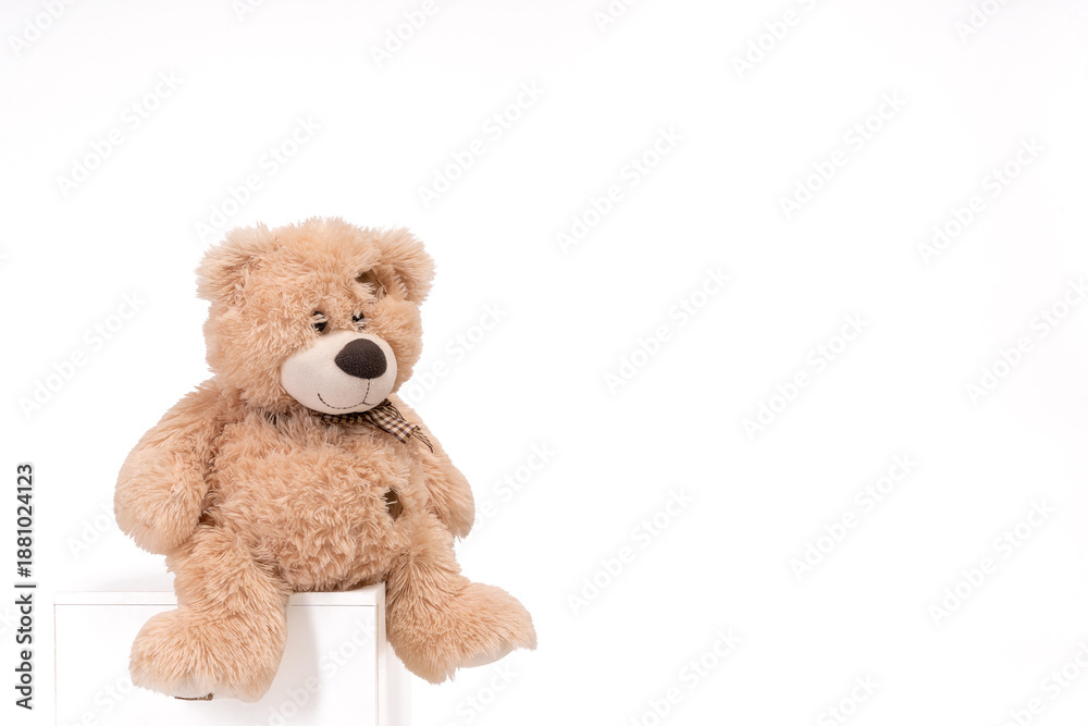 Fototapeta premium Cute brown teddy bear sitting on a white cube against a clean white background with generous copy space. The soft plush texture and warm neutral tones create a cozy, comforting childhood atmosphere