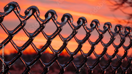 A close-up shot of a chain-link fence encased in a thick, clear layer of ice following a freezing rain storm. The background features a dramatic and vibrant sunset with deep orange, pink, and red hues