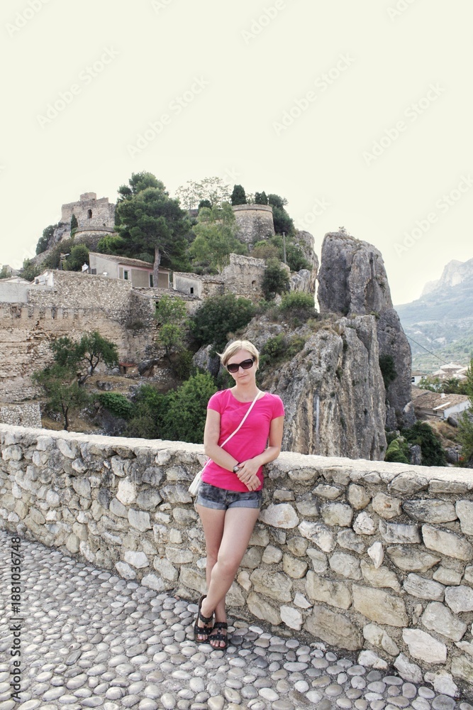Fototapeta premium Female tourist posing for a photo on a stone wall overlooking the historic Guadalest castle ruins and mountain village architecture in Spain on a sunny day