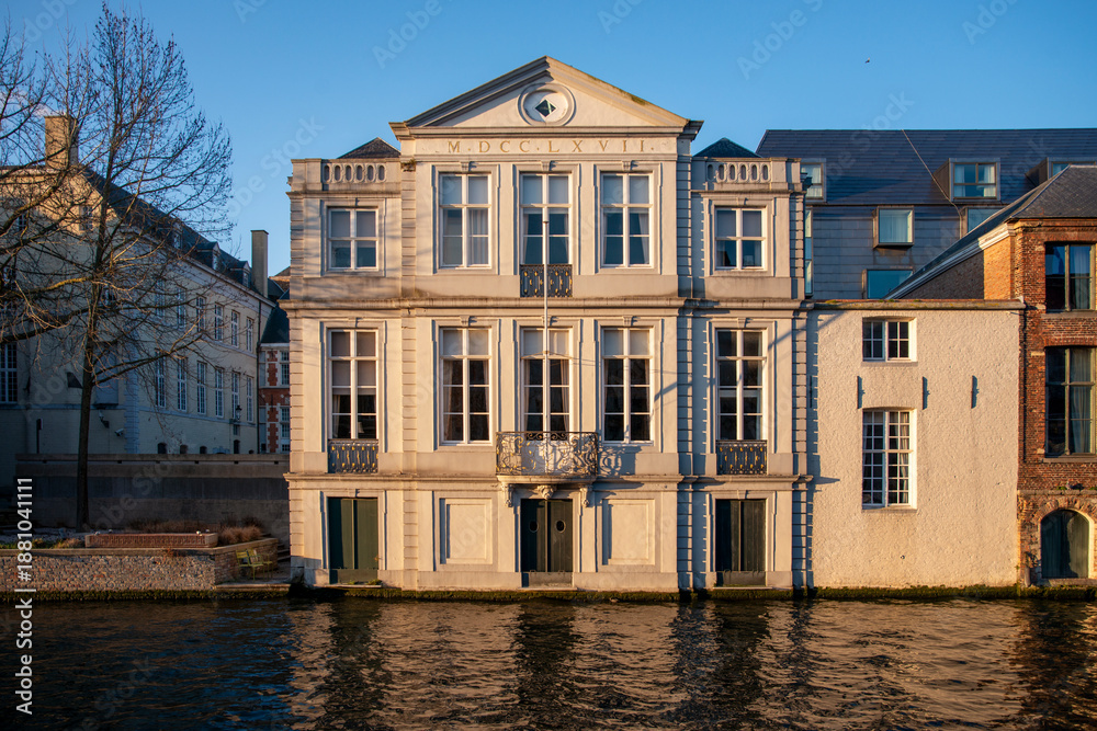 Obraz premium Elegant neoclassical canal house in Bruges with Roman numeral pediment and tall white windows reflecting warm golden hour light on the water