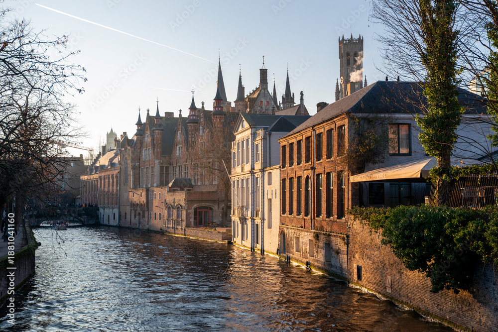 Fototapeta premium Golden hour view of Bruges canal with medieval brick buildings and gothic spires reflecting in rippling water, historic old town cityscape in Belgium