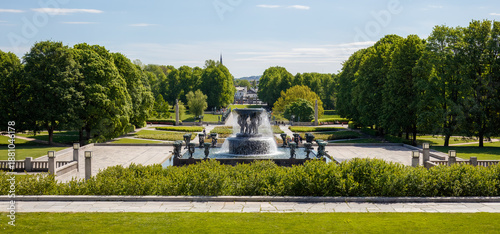 Busy city quiet moment in Frognerpark with fountain surrounded by lush greenery, pathways and serene landscape during a sunny day