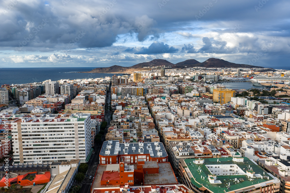 Obraz premium Aerial cityscape of Las Palmas de Gran Canaria shows white and pastel blocks, La Isleta twin peaks, port basin, cruise ships, winter light, and dramatic clouds.