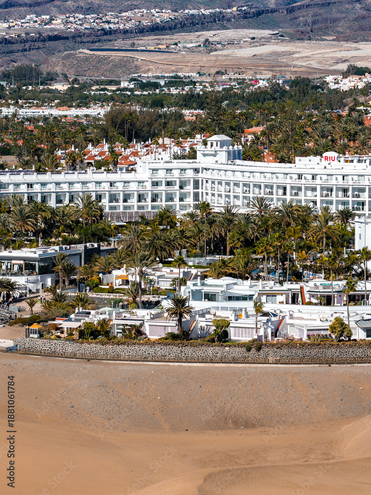 Fototapeta premium Aerial view of Maspalomas Dunes in Gran Canaria with the white RIU hotel, beachfront hotels, villas, pools, stone seawall, palm groves, and distant hills in bright midday light.