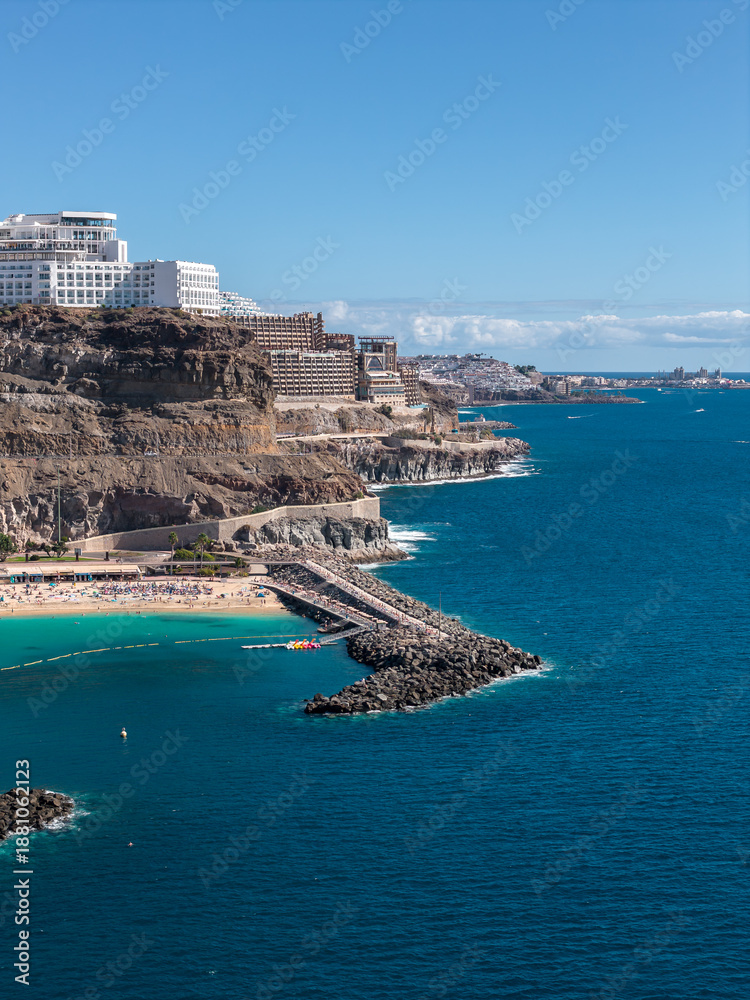 Fototapeta premium High angle view of Amadores Beach near Puerto Rico de Gran Canaria shows rugged cliffs, stepped hotels, a curved breakwater, busy sand, clear sky, and calm Atlantic.