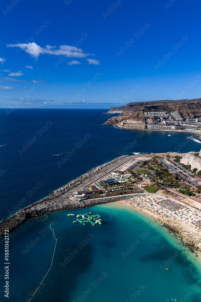 Fototapeta premium Aerial view shows Amadores Beach, Gran Canaria, sunbathers, umbrellas, curving breakwater, marina, inflatable park, boats, terraced hotels, and distant Mount Teide.
