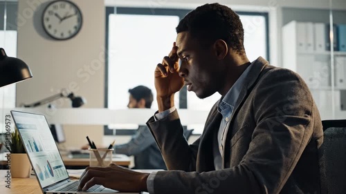 An African American businessman sits at his modern office desk, looking frustrated and holding his head in a moment of stress. His laptop displays various charts and graphs, indicating a demanding