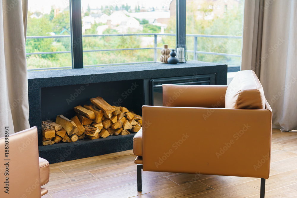 Fototapeta premium Stylish modern living room interior with fireplace, firewood and comfortable leather brown armchairs on large window background. Warm cozy home atmosphere, contemporary design and natural daylight.