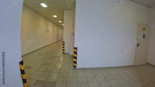 Long, narrow corridor with textured white walls, tiled floor, and fluorescent lighting. Backstage. This creates sense of anxiety and disorientation, confinement, isolation. Slow, forward movement. Not