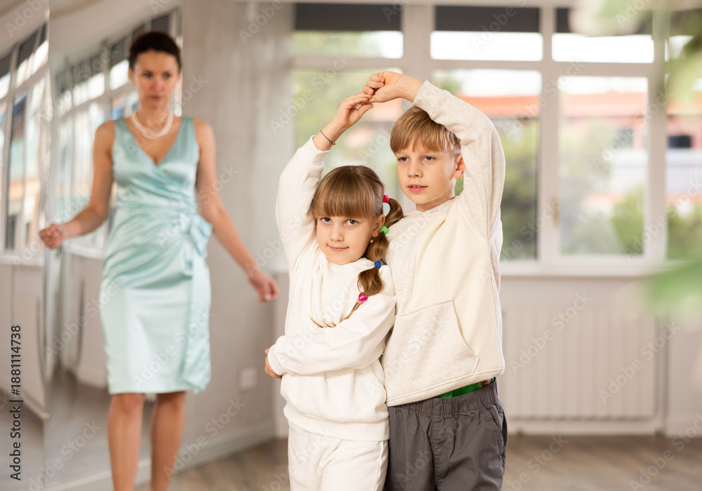 Obraz premium Couple of positive interested preteen dancers, boy and girl preparing for performance, rehearsing ballroom dance in spacious classroom while female teacher observing from background