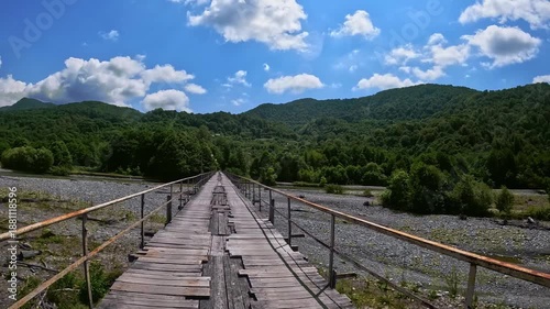 Worn, old wooden suspension bridge with missing planks and metal railings crosses wide riverbed. Fictional character walks across bridge. Natural landscape. Slow Motion Video, Not AI