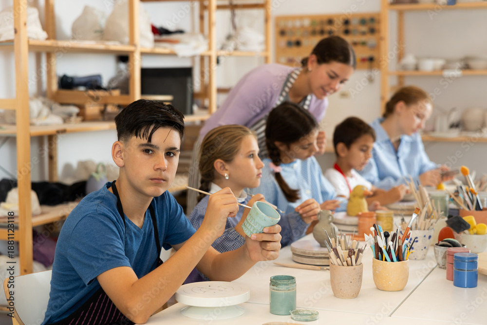 Obraz premium Teenager paints a clay mug with brush at a workshop with a teacher. Boy at a ceramics master class works at a table in an art studio