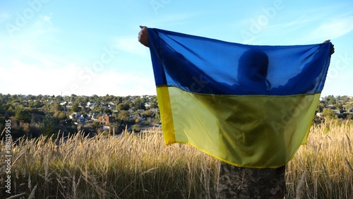 Military man in uniform stands with raised over head flag of Ukraine at countryside. Male ukrainian army soldier with lifted national banner in honor of victory against russian aggression. End of war