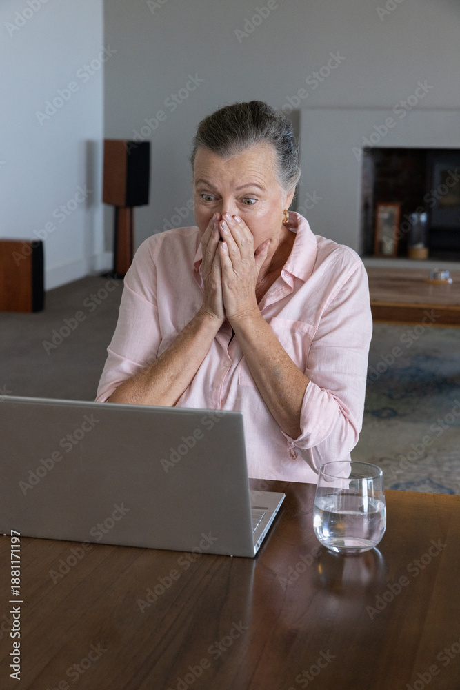 Fototapeta premium Senior woman sitting at living-room table in pink shirt using open laptop, covering mouth, glass