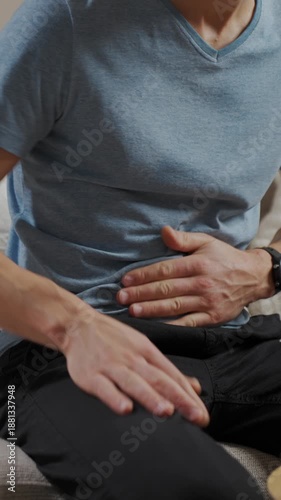 Man sitting on sofa in living room holding stomach and suffering from abdominal pain or cramps. Person suffering from health issues, digestion problems or food poisoning at home