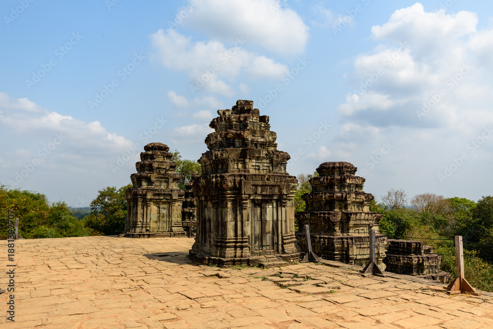 Obraz premium Three weathered sandstone towers rise from the sunlit terrace of Phnom Bakheng temple near Siem Reap, Cambodia. The ancient structures stand against a backdrop of green treetops and a sky dotted with