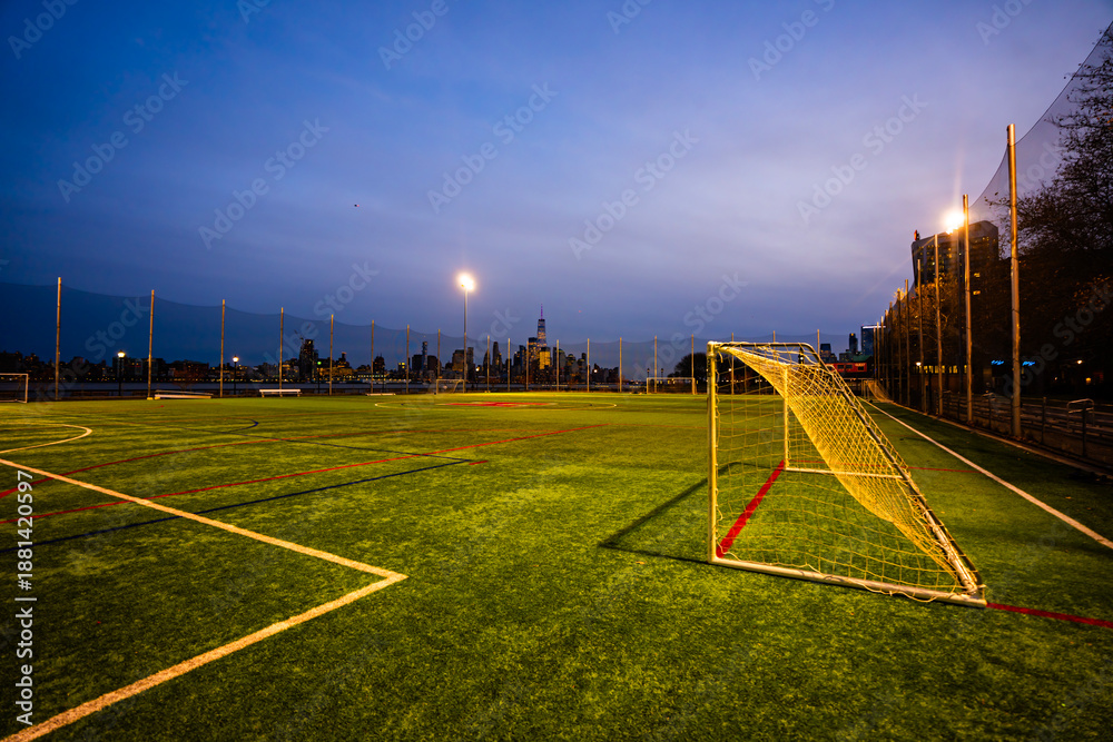 Fototapeta premium Empty stadium field with football lines and Manhattan skyline in the background. Soccer stadium in NYC with green turf and city lights at dusk. Urban football stadium overlooking Manhattan.