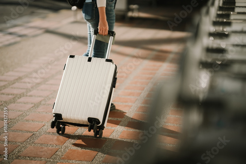 Fotografie Close-up of a young Asian woman holding a passport while pulling a rolling suitcase at an airport terminal, representing international travel, mobility, and modern journey lifestyle