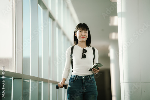 Free Download Young Asian woman standing in a modern airport terminal, using a smartphone while holding a passport, with travel luggage nearby, preparing for a journey in a bright, contemporary space. - GFXLoad