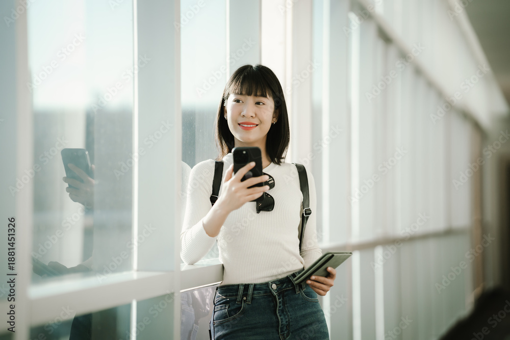 Fototapeta premium Young Asian woman standing in a modern airport terminal, using a smartphone while holding a passport, with travel luggage nearby, preparing for a journey in a bright, contemporary space.