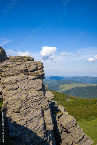 A dramatic vertical rock formation rises sharply above rolling forested hills and distant valleys under a vivid blue sky with scattered clouds at Mont de Gerbier de Jonc.