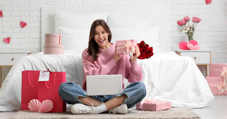 Beautiful young woman with laptop, gifts and shopping bags sitting on floor near bed in bedroom. Valentine's Day celebration