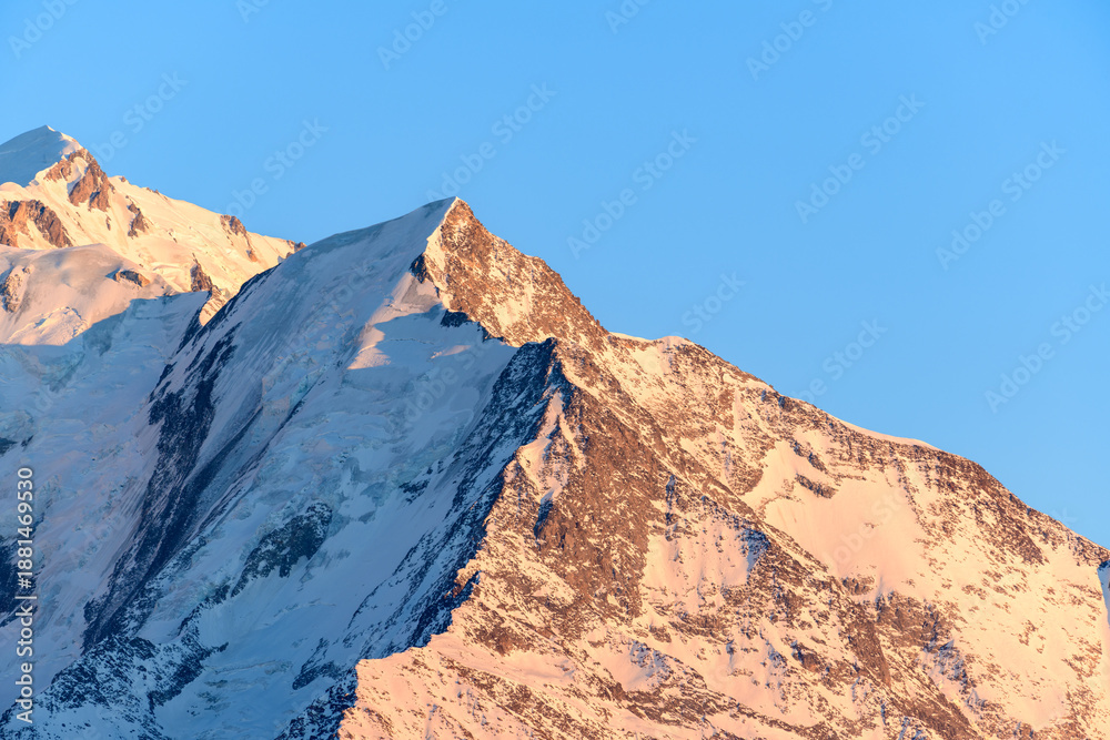 Fototapeta premium Golden sunlight illuminates the snowy, rugged slopes of Aiguille de Bionnassay, revealing crisp textures and sharp contrasts beneath a clear blue sky in the French Alps.
