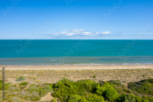 Wallpaper Mural Expansive view of a quiet sandy beach bordered by wild green shrubs and calm turquoise sea under a bright blue sky, evoking a sense of peace and open space. Torontodigital.ca