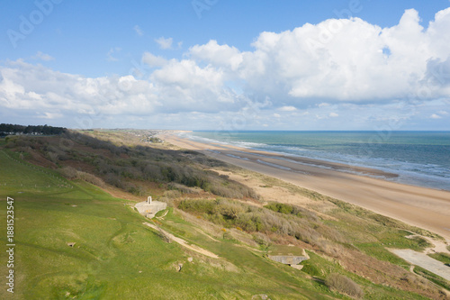 Wide aerial perspective of Omaha Beach in Normandy, France, showing grassy bluffs, sandy shoreline, and the blue expanse of the Atlantic Ocean beneath a sky filled with scattered clouds. The landscape