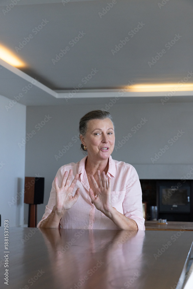 Fototapeta premium Mature adult woman wearing pink blouse speaking gesturing at large wooden table in modern home