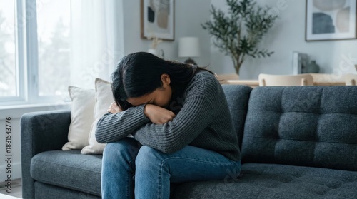 Sad Southeast Asian adult woman sitting alone on a gray sofa indoors, leaning forward with head in hands expressing anxiety and depression in a bright living room, mental health concept.
