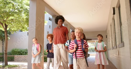Teacher with children guiding along walkway with columns backpacks as blonde boy stepping to lead